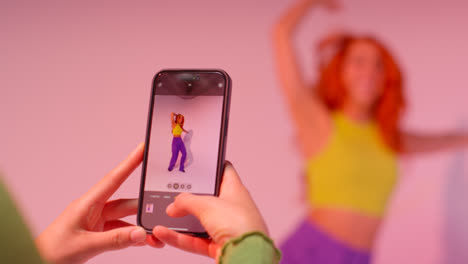 Studio-Shot-Of-Woman-Taking-Photo-Of-Friend-Dancing-On-Mobile-Phone-Against-Pink-Background-4